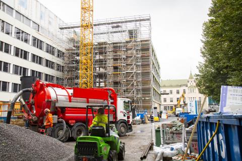 Valkoisen ulkoa valmiin talon ja rakennustelineiden peitossa olevan uudisosan edessä on punainen säiliauto, soraa ja pieni työkone kuljettajineen.