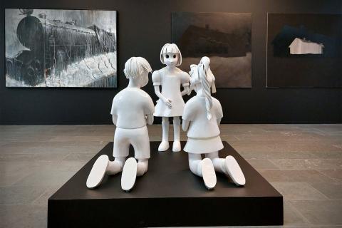 Edessä valkoinen veistos, joka esittää kolmea lapsihahmoa. Taustalla tummia maalauksia. 