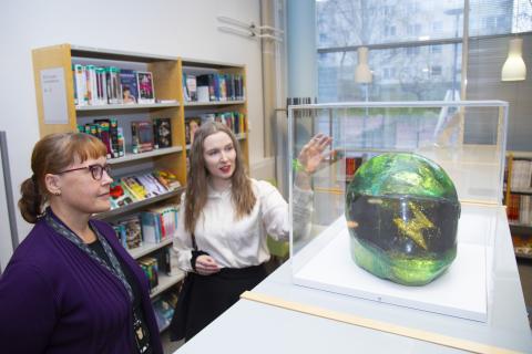 Kaksi henkilöä katsoo vitriinin sisällä olevaa kypärää. Taustalla on kirjahyllyjä.