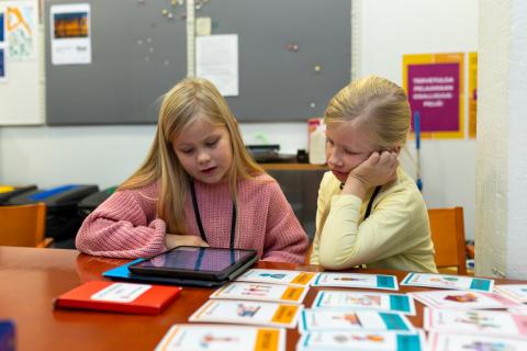 Kaksi tyttöä istuu pöydän ääressä ja pelaa iPadilla osallisuuspeliä.