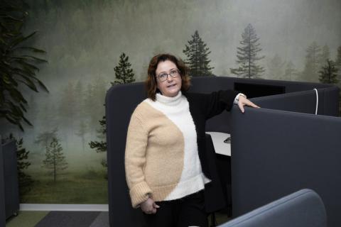 Silmälasipäinen henkilö seisoo ja katsoo kameraan. Taustalla olevassa seinässä on metsäkuvioinen valokuvatapetti.