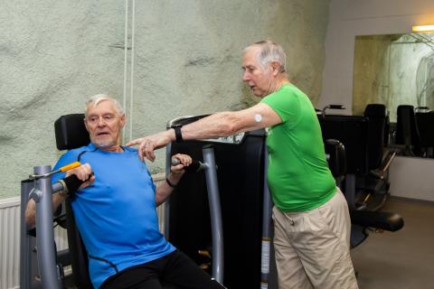 Kaksi vanhempaa miestä kuntosalilaitetta käyttämässä. Toinen opastaa toista.