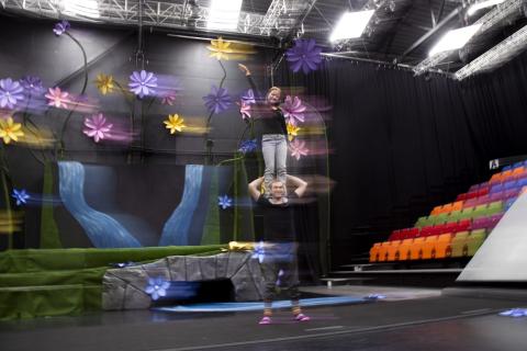 Ihminen seisoo toisen ihmisen hartioilla tummassa esitystilassa, jossa on värikkäitä kukkia taustalla. Tilassa on myös värikkäitä katsomotuoleja.