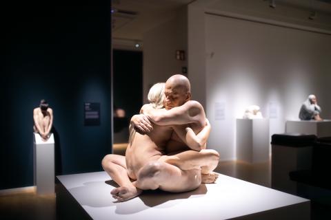 Hyperrealistista taidetta näyttelysalissa, etualalla veistos, jossa alastomat mies- ja naishahmo halaavat toisiaan.
