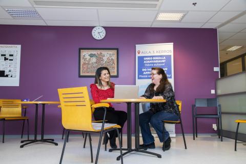 Kaksi henkilöä istuu huoneessa pöydän ääressä tietokoneella jutellen keskenään.