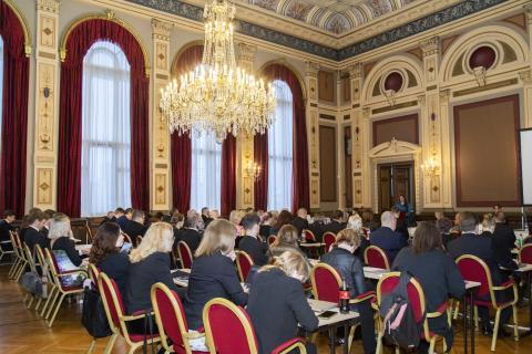 The Council sits in its red chairs in the banquet hall under a crystal lamp.