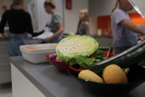 Kaali, nauriita, kesäkurpitsaa ja muita kasviksia, taustalla ihmisiä kokkaamassa.