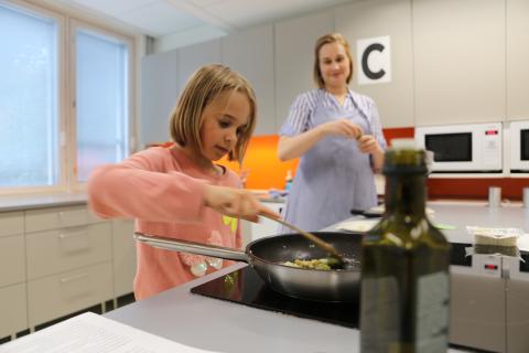 Lapsi lisää mausteita paistinpannulle kotitalousluokan keittiössä, äiti taustalla.