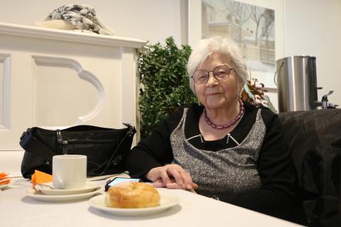 Henkilö hymyilee pöydän ääressä, kahvia ja pullaa.