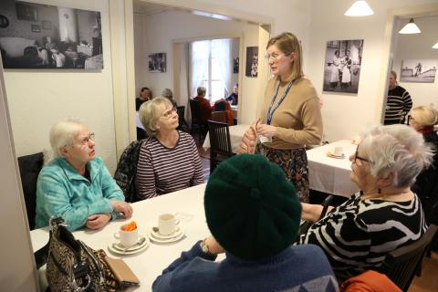 Neljä henkilöä istuu kahvilan pöydän ääressä, yksi henkilö seisoo.