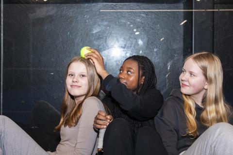 Three youngster sits on the padel floor and one of them holds a tennis ball above another&#039;s head.