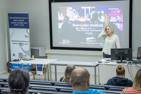 Kuvan etualalla on henkilöitä kuuntelemassa luentoa. Puhujan taustalla on heijastettu diakuva aiheesta.