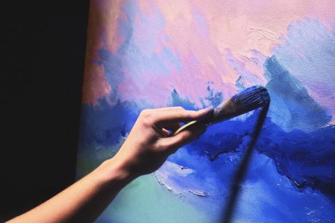Käsi pitelee pensseliä, joka maalaa sinisiä ja vaaleanpunaisia sävyjä kankaalle.
