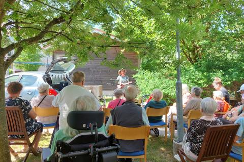 Ryhmä ikäihmisiä istuu ja laulaa kesäisen vihreän puulehvistön alla.