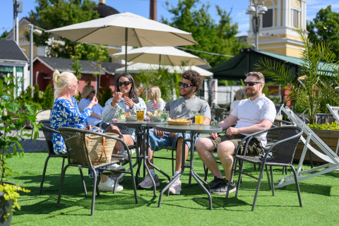 Neljä ihmistä nauttimassa aurinkoisesta päivästä terassipöytien ääressä ruoan ja juoman kera.
