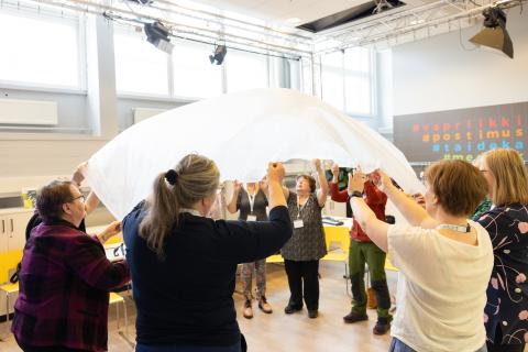 Naiset seisovat piirissä ja pitävät kiinni pyöreän muotoisesta valkoisesta harsosta.