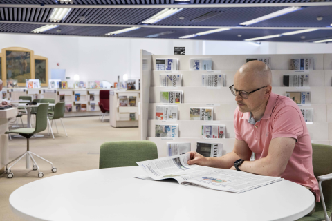 Henkilö lukee lehteä kirjaston lehtilukusalin pöydän ääressä. Takana on lehtihyllyjä.