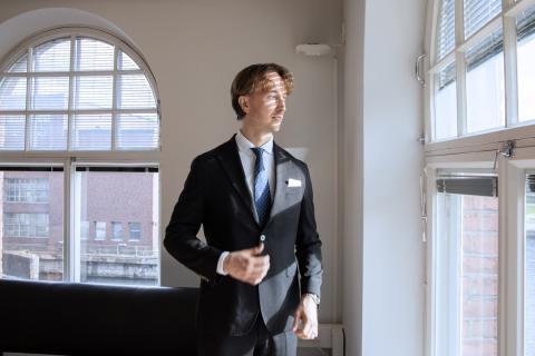 Pormestari katselee rakennuksen kulmassa sijaitsevan työhuoneensa ikkunasta ulos. Takana on toinen kaareva ikkuna, josta näkyy punatiilinen rakennus ja hieman Tammerkoskea.