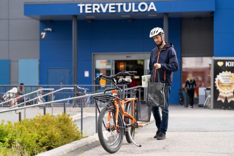 Henkilö tulossa kaupasta, laittaa ostoksia pitkäperäisen tavarapyörän koriin.