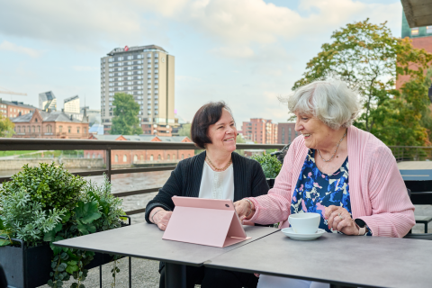 Kaksi ikääntynyttä henkilöä istuu pöydän ääressä edessään tablet-laite ja kahvikuppi.