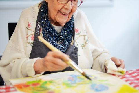 Iäkäs nainen iloinen ilme kasvoillaan maalaa isolla pensselillä värikästä kuvaa.