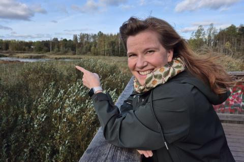 Nainen hymyilee iloisena Iidesjärven lintutornissa ja osoittaa järvelle. 