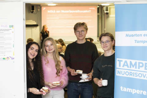 Neljä henkilöä seisoo kakkukahvit kädessään ovensuussa. Vieressä kyltti ja takana suuri näyttö, joissa on tekstejä Tampereen nuorisovaltuustosta.