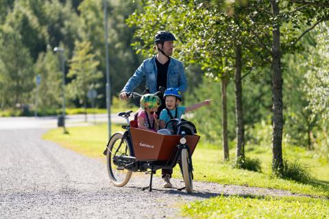 Kaksi lasta istuu tyytyväisinä tavarapyörän etulaatikossa, mies pitää pyörää valmiina lähtemään liikkeelle. 