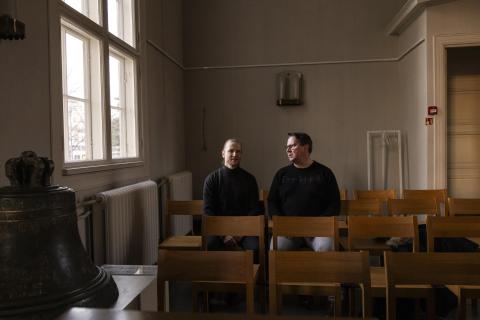 Kaksi tummiin pukeutunutta henkilöä istuu puutuoleilla kirkkosalin perällä. Vasemmalla sivussa on vanha kirkonkello.