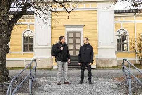 Kaksi tummiin talvivaatteisin pukeutunutta henkilöä seisoo ja juttelee keltaisen kirkon edessä.