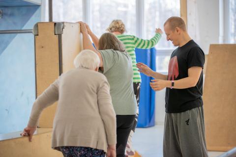 Hymyilevä mies ohjaa ikäihmisiä liikuntasalissa.