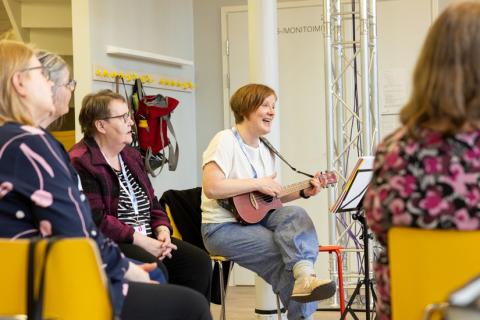 Nainen soittaa kitaraa ja laulaa neljän muun naisen kanssa.