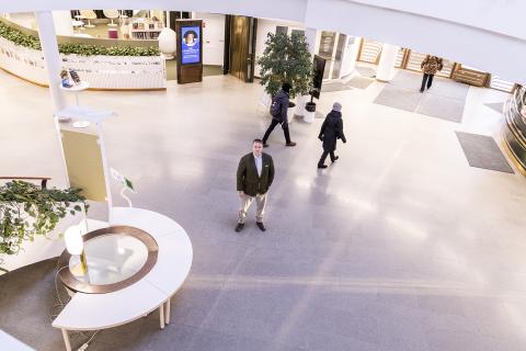 A person is standing in a spacious lobby, looking up toward the camera. People are walking in the background.