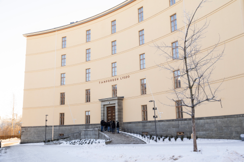 A pale yellow building with the words “Tampereen lyseo” on it. In front of it is a snowy pavement and a single tree.