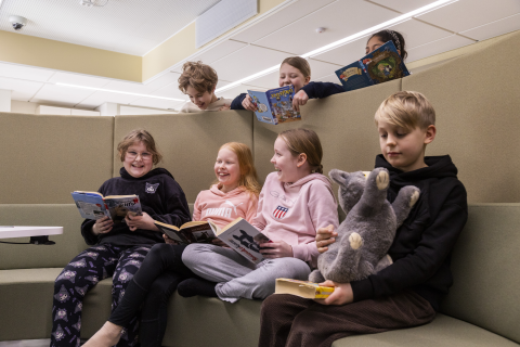 Koulun oppilaita istuu kirjojen ja pehmolelun kanssa ja nauraa. Osa oppilaista kurkkii ylhäältä sermien takaa.