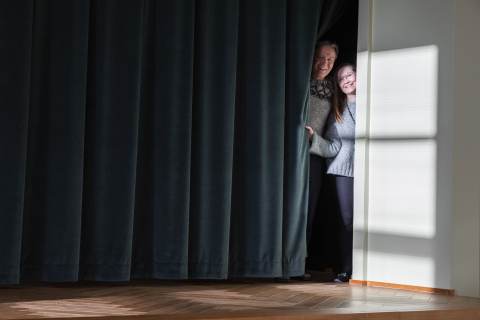 Kaksi henkilöä kurkistaa esiripun sivusta.