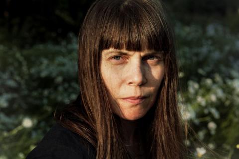 Hulda Huima katsoo kameraan vakavalla ilmeellä. Taustalla näkyy sumeasti valkoisia kukkia.