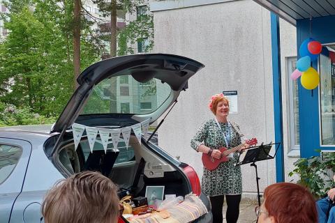 Nainen soittaa ukuleleä auton avonaisen tavaratilan vieressä.