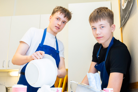 Kaksi henkilöä katsoo kameraan. Toinen on juuri tyhjentämässä kulhoa pöydällä olevaan astiaan.