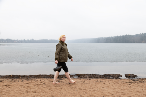 Henkilö kävelee hiekkarannalla paksussa takissa, housujen lahkeet käärittyinä ylös ja pitelee kädessään kenkiä.