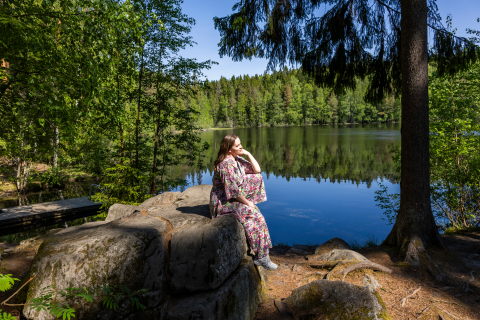 Henkilö istuu suurella kivellä pienen järven rannalla. Ympäröivä metsä heijastuu järven pinnasta.