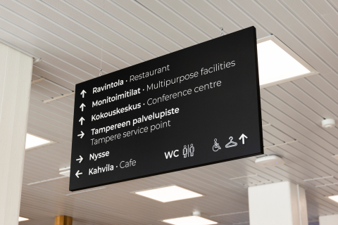 Musta opastekyltti roikkuu katossa. Tekstinä suomeksi ja englanniksi: ravintola, monitoimitilat, kokouskeskus, Tampereen palvelupiste, Nysse ja kahvila sekä wc.