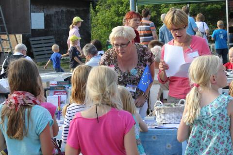 Kaksi kaupungin edustajaa ulkona kesätapahtumassa esittelypöydän ääressä. Vaaleatukkainen kumartuu kohti pöydän toisella puolella olevaa lapsiryhmää.