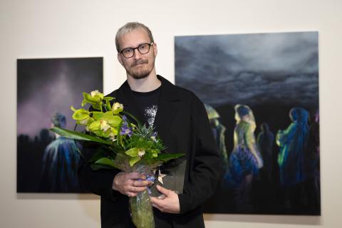Vuoden nuori taiteilija 2026 Roope Itälinna seisoo maalausten edessä kukkakimppu kädessä.