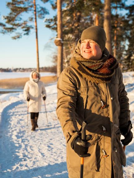 Pariskunta sauvakävelee lumisessa maisemassa auringon paisteessa.