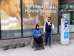 Pyörätuolissa istuu liikuntavammainen nainen ja hänen vierellään nakövammainen mies, joka pitelee sokean valkoista keppiä. He ovat kadun varrella lähdössä liikenteeseen.